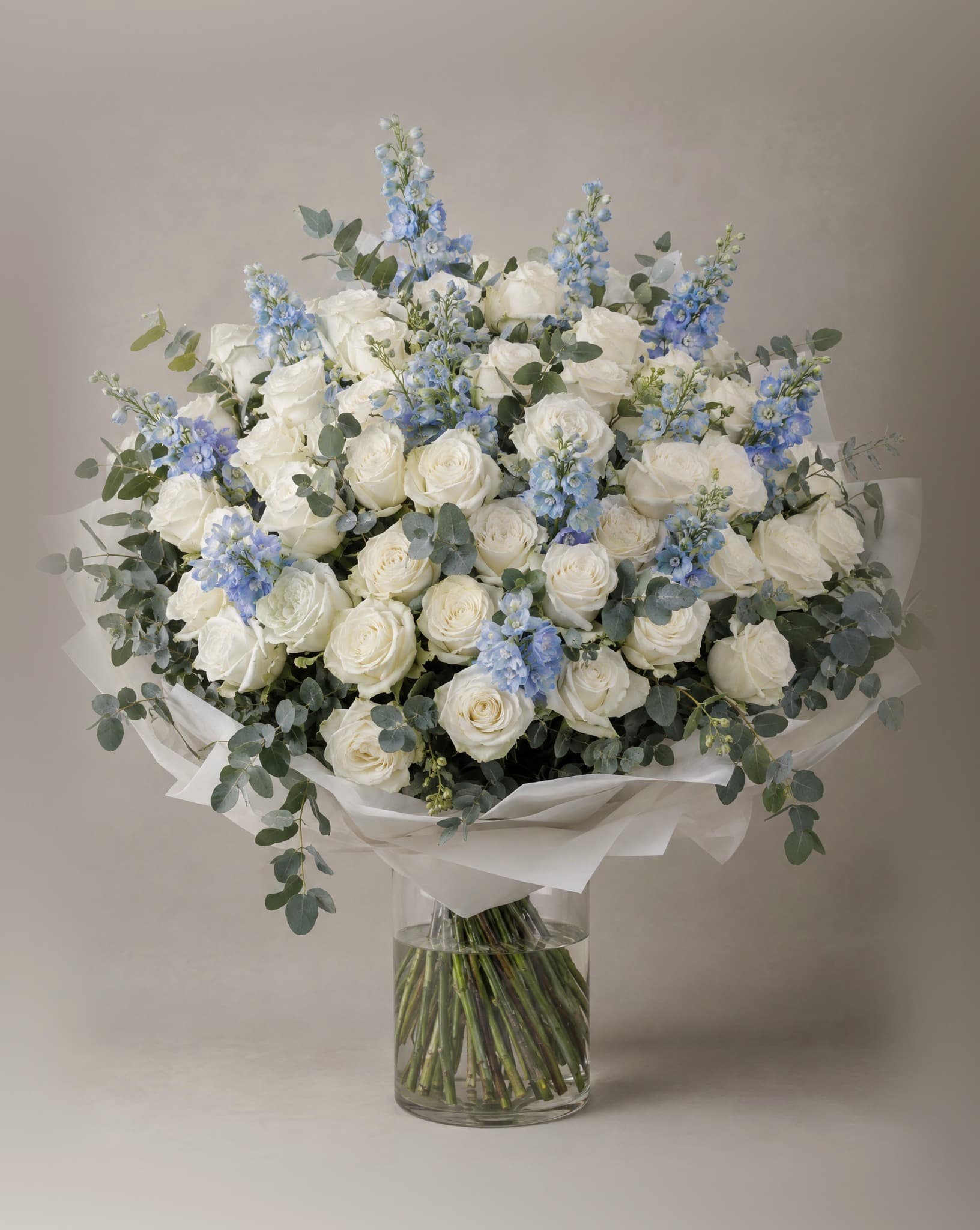 One hundred white roses with blue delphinium and eucalyptus in a wide domed bouquet in a clear glass vase