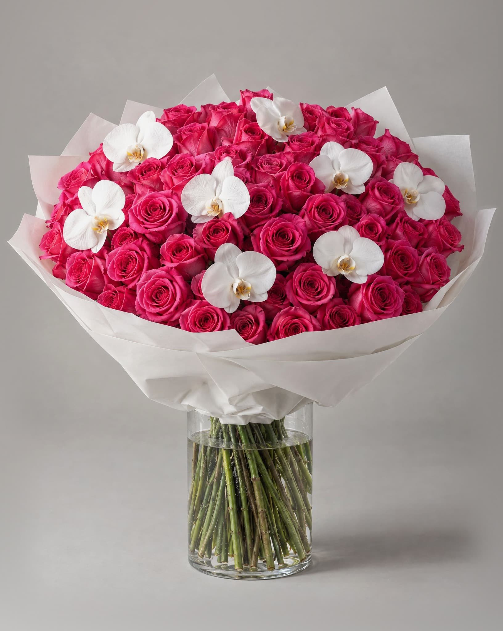 100 hot pink roses with 8 white phalaenopsis orchids wrapped in white paper in a clear glass vase